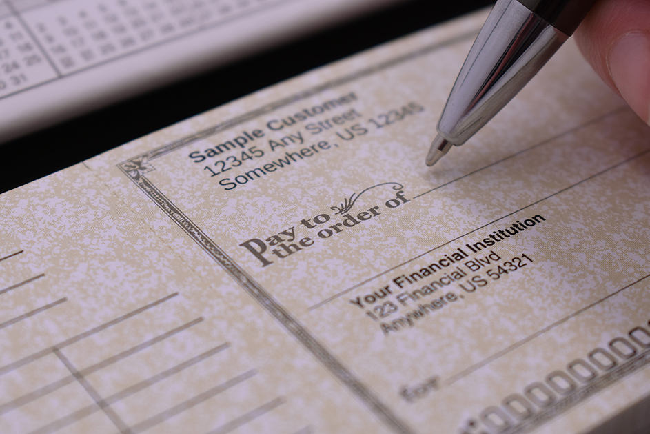 A close-up shot of a hand using a silver pen to fill out a personalized sample check with a parchment-style background. The image highlights the "Pay to the order of" line and customizable account details, ideal for showcasing custom banking stationery.